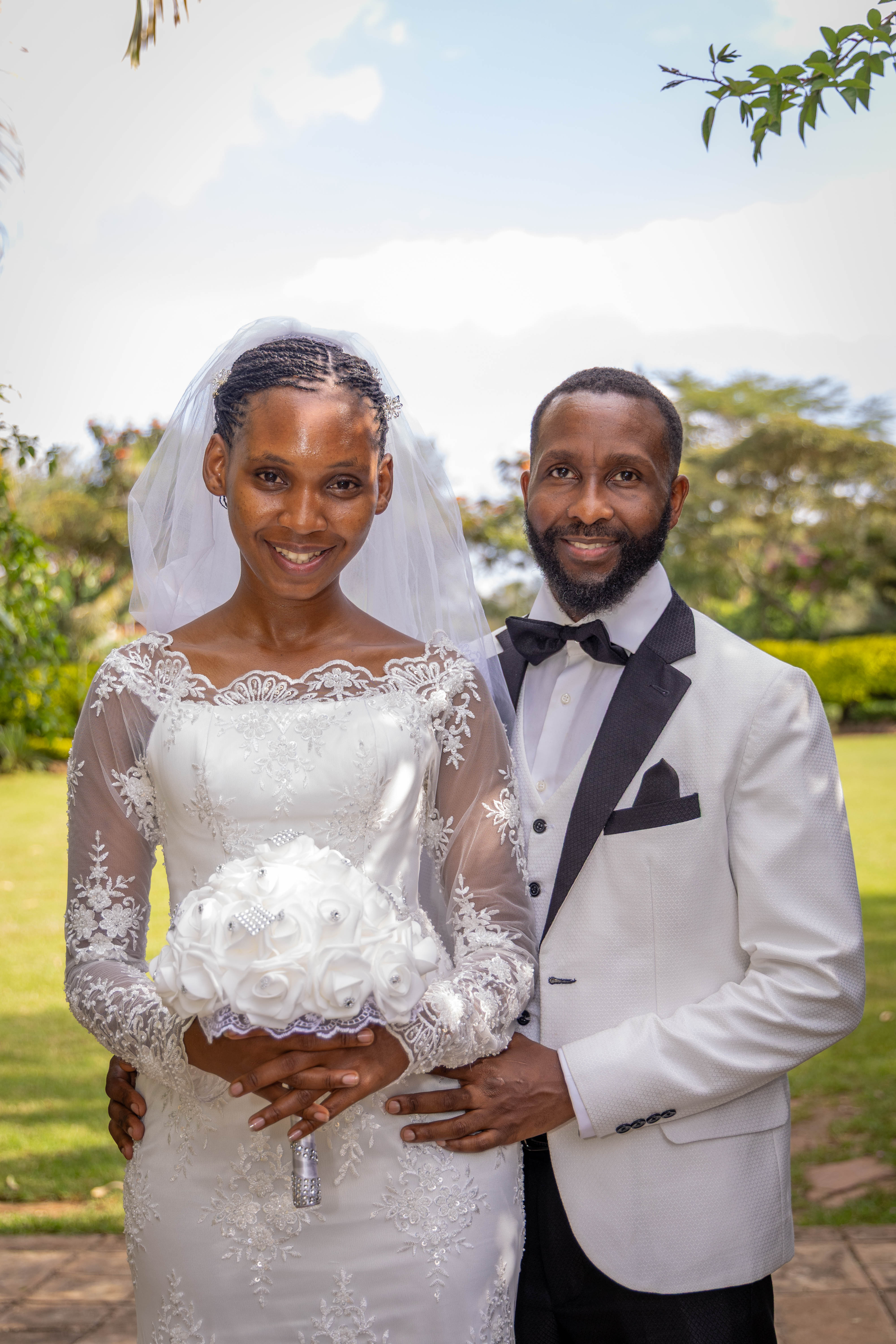 The bride holds a white bouquet as the groom stands close beside her in a garden