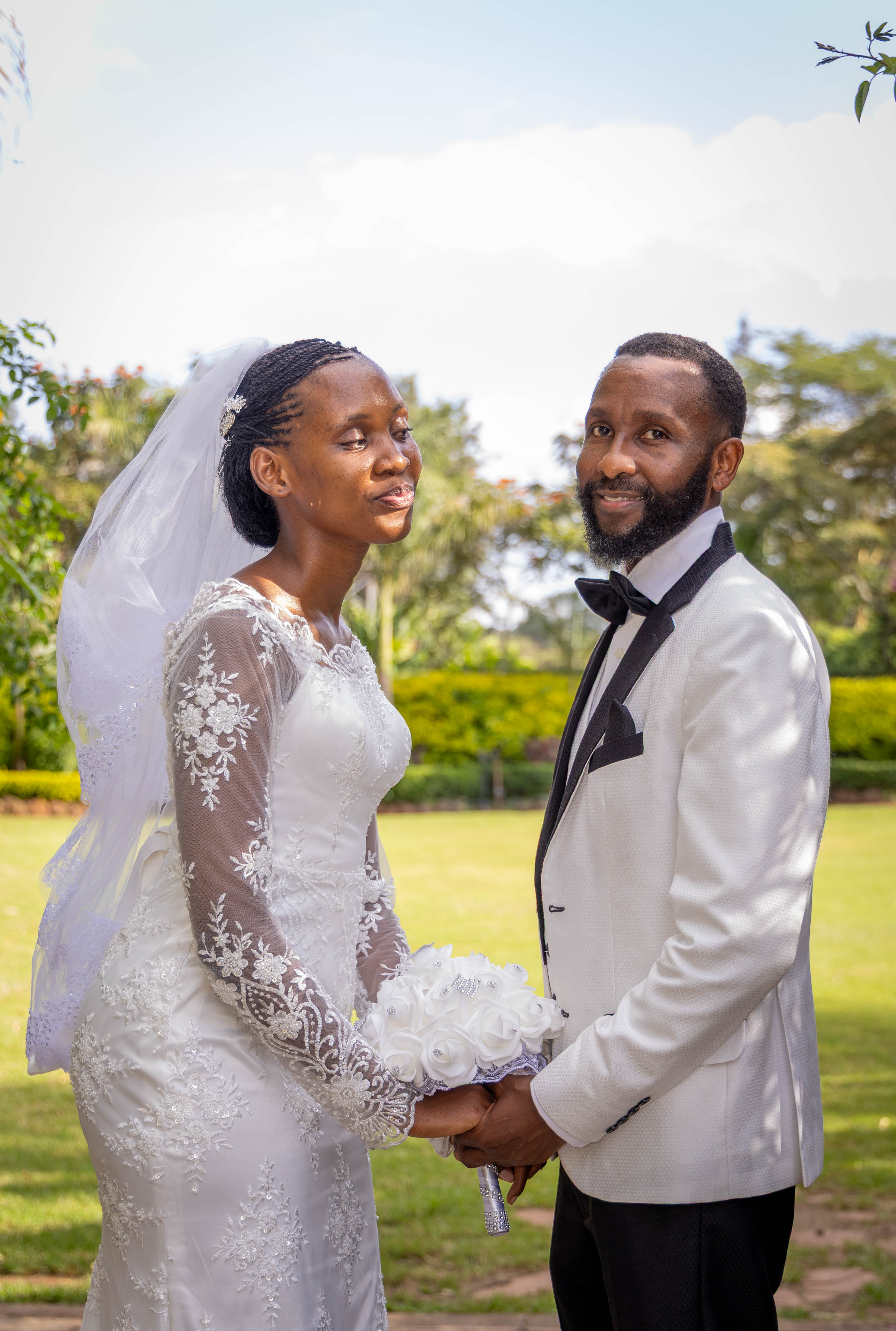 The bride and groom face each other on a lawn, holding hands