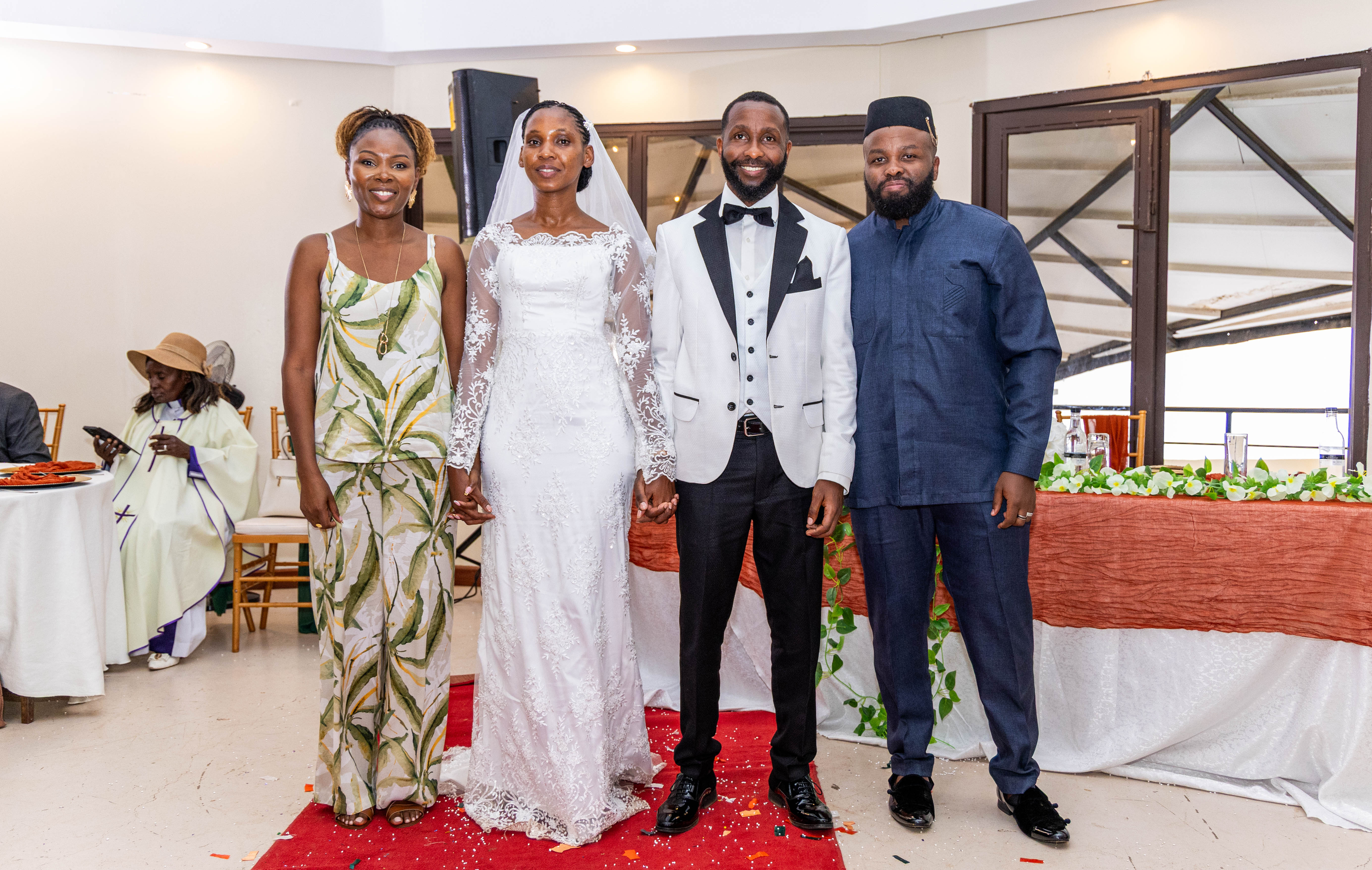 The couple stands on a red carpet with two guests at the reception venue