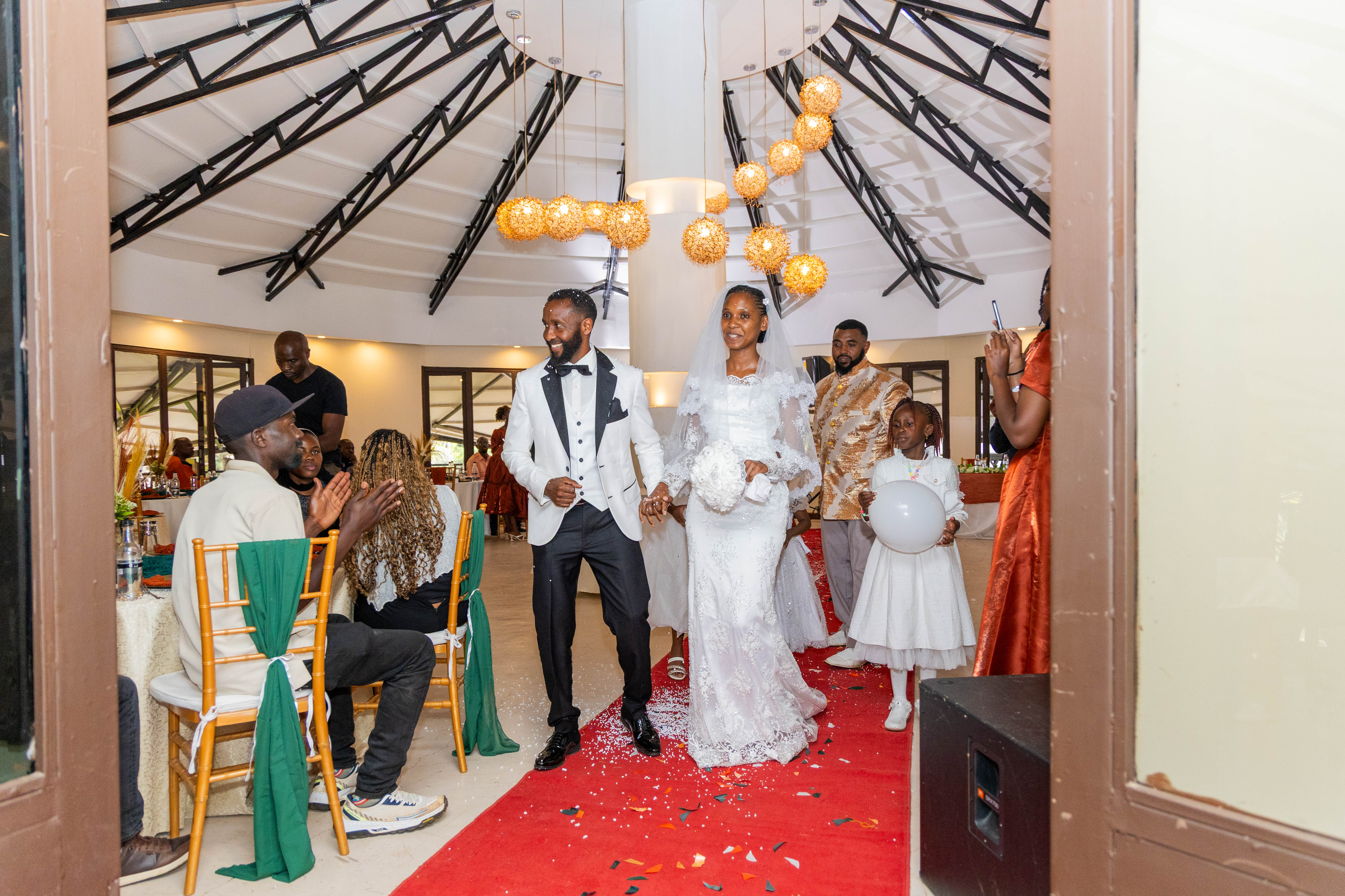 The bride and groom walk down a red carpet into the reception hall as guests cheer