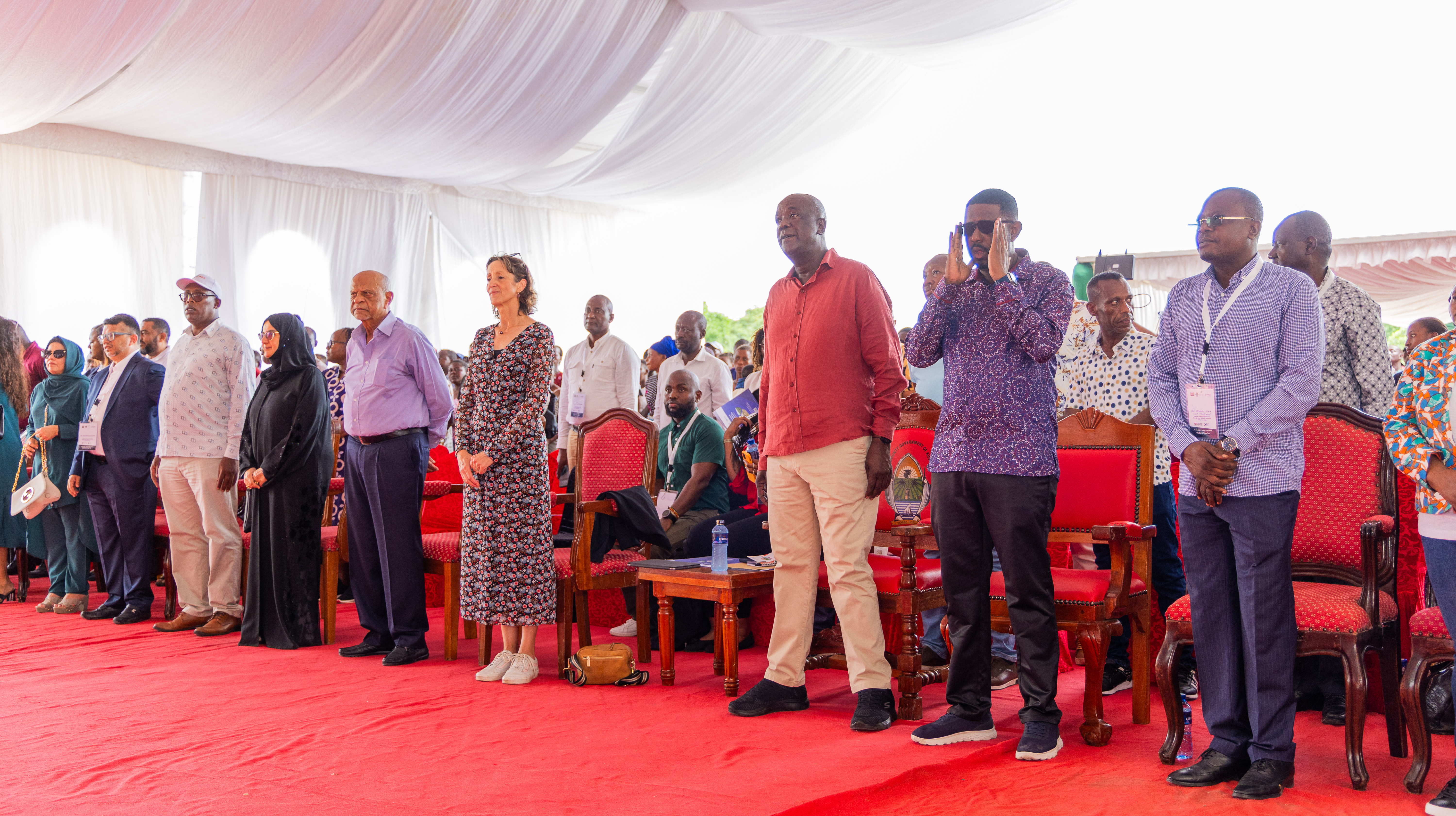 Guests stand in rows on a red carpet under a white tent