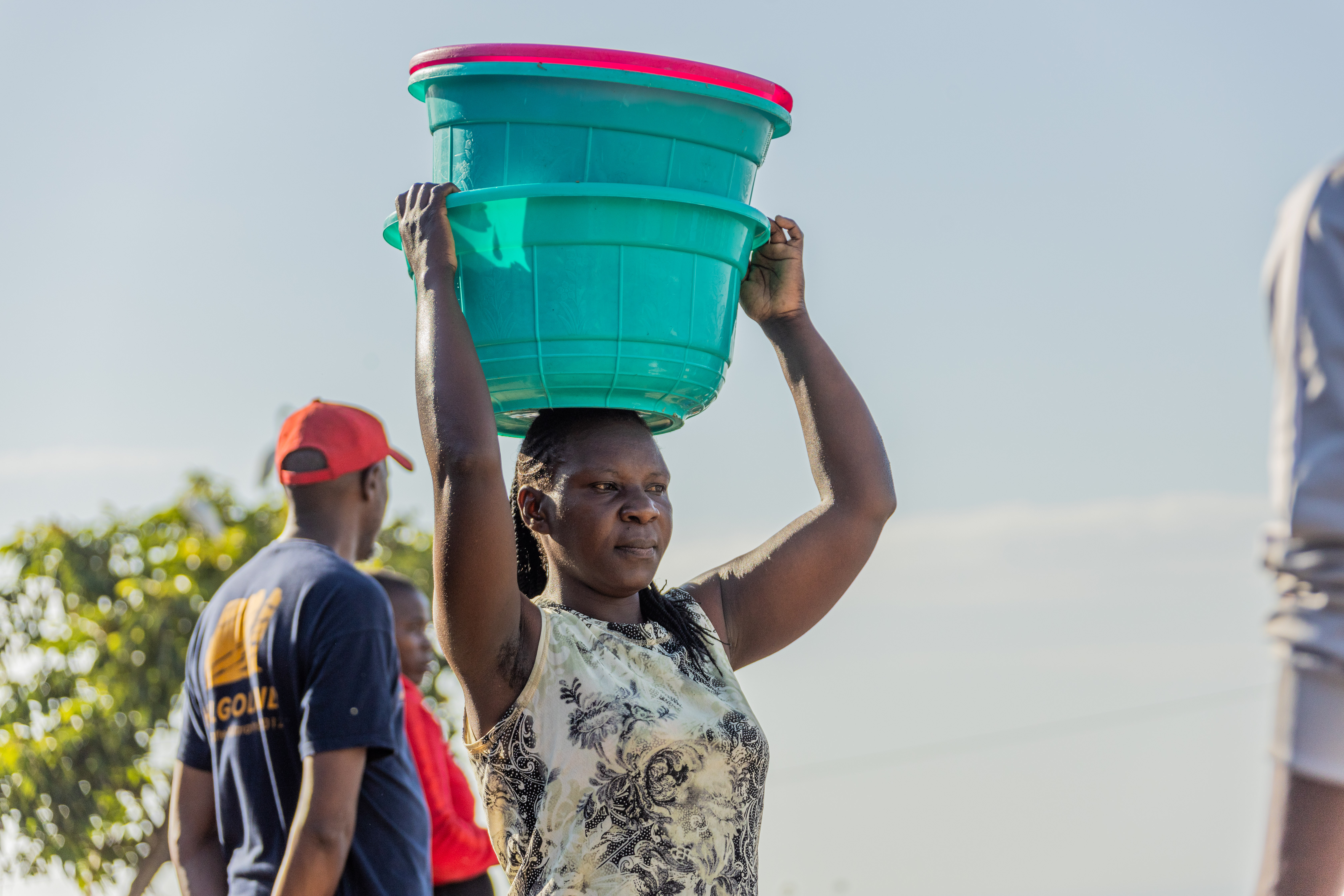 A woman carries two stacked turquoise basins on her head against a pale sky