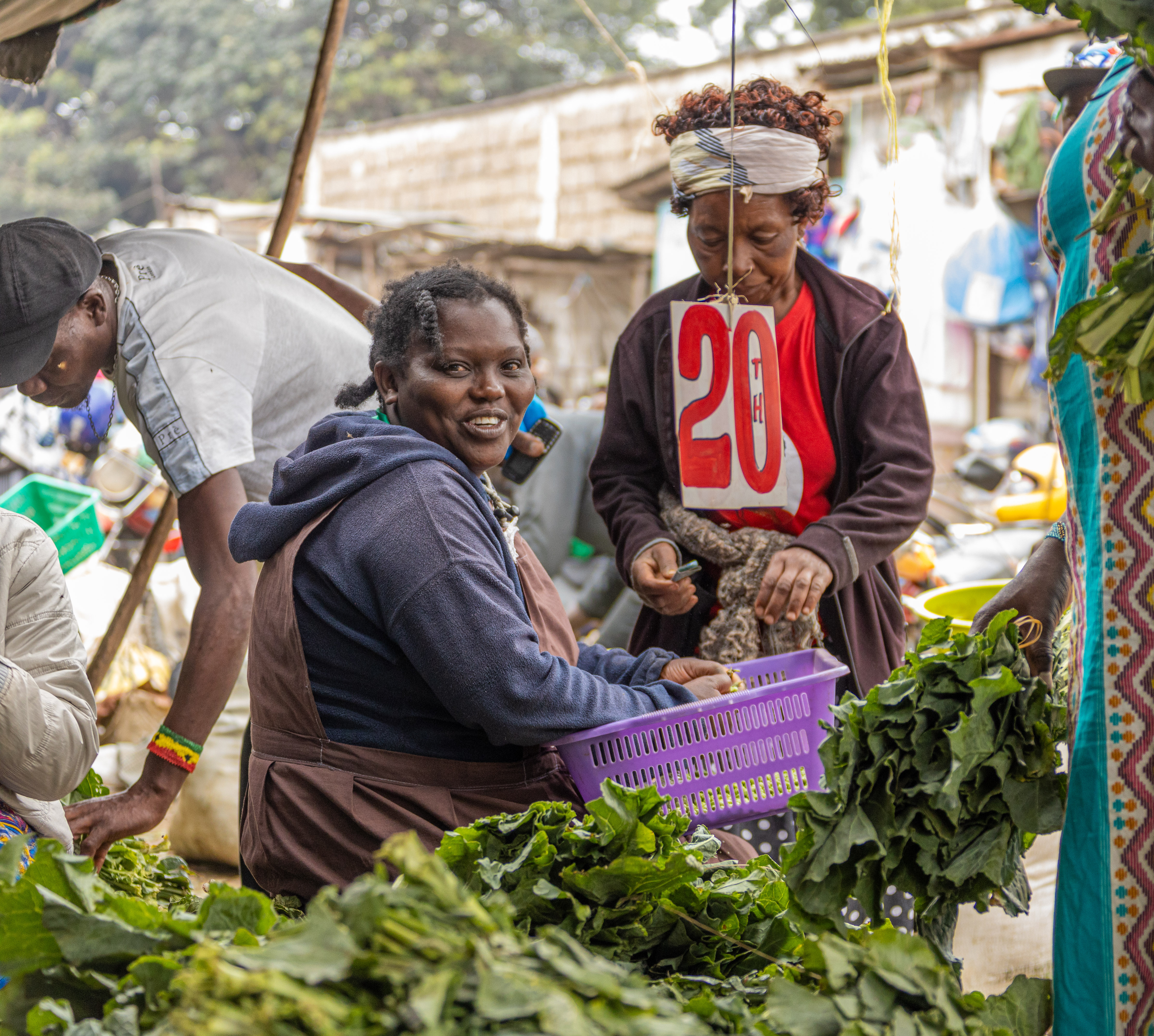 A smiling vegetable seller works among bundles of greens at a market