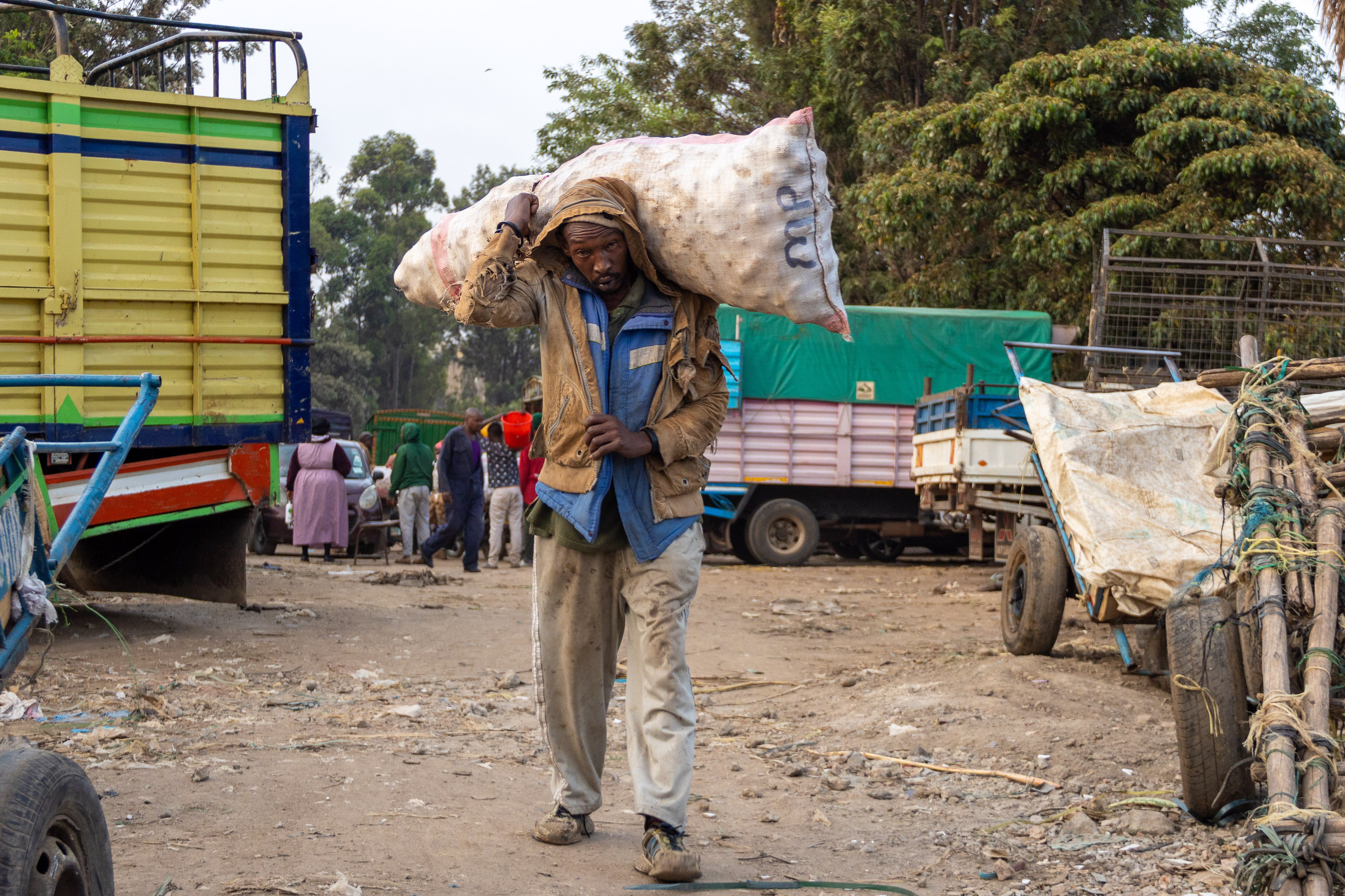 A porter carries a heavy sack on his shoulders between parked lorries