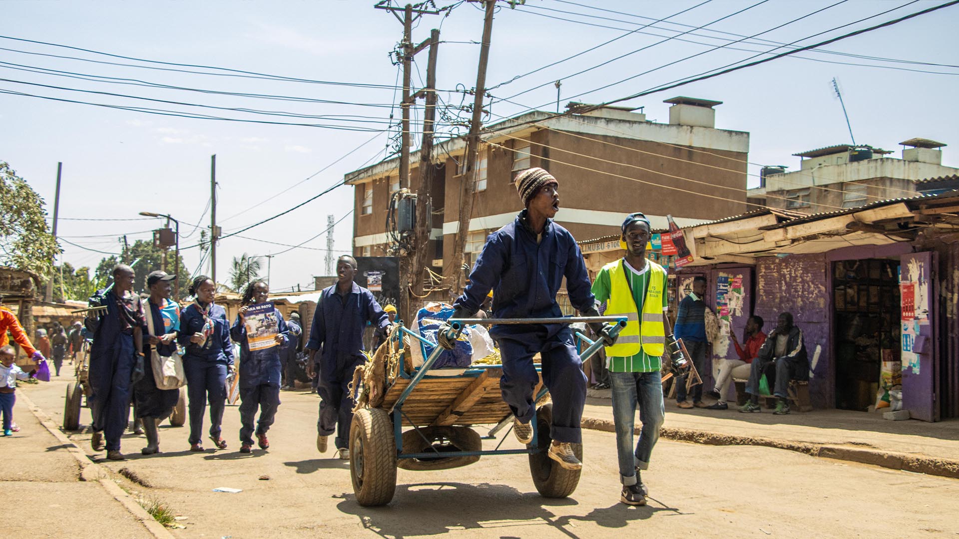 Waste collectors in navy overalls push a loaded handcart down a busy street