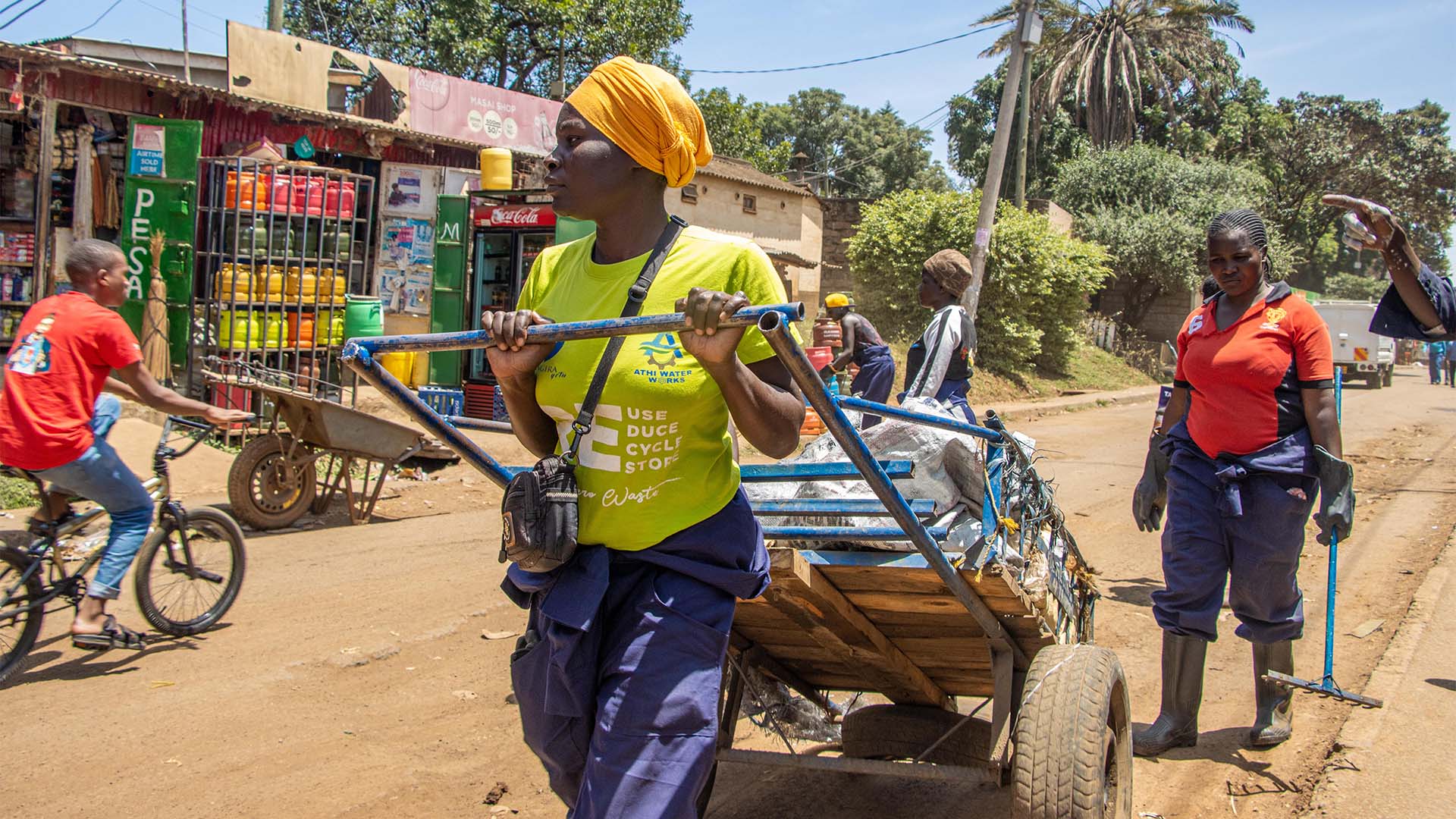A woman in a yellow headscarf pulls a handcart past roadside shops
