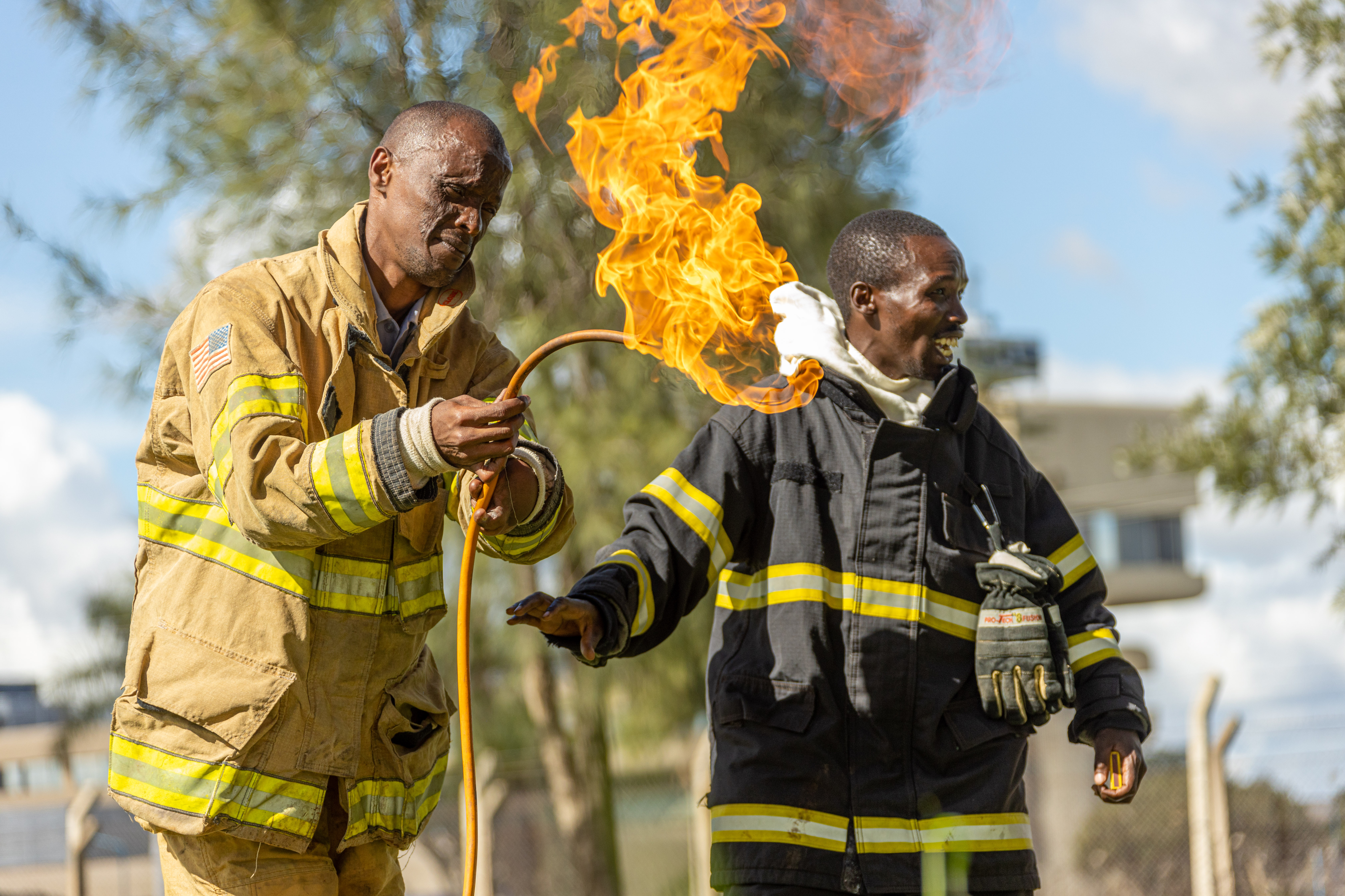 A trainer guides a trainee in turnout gear through a live flame exercise