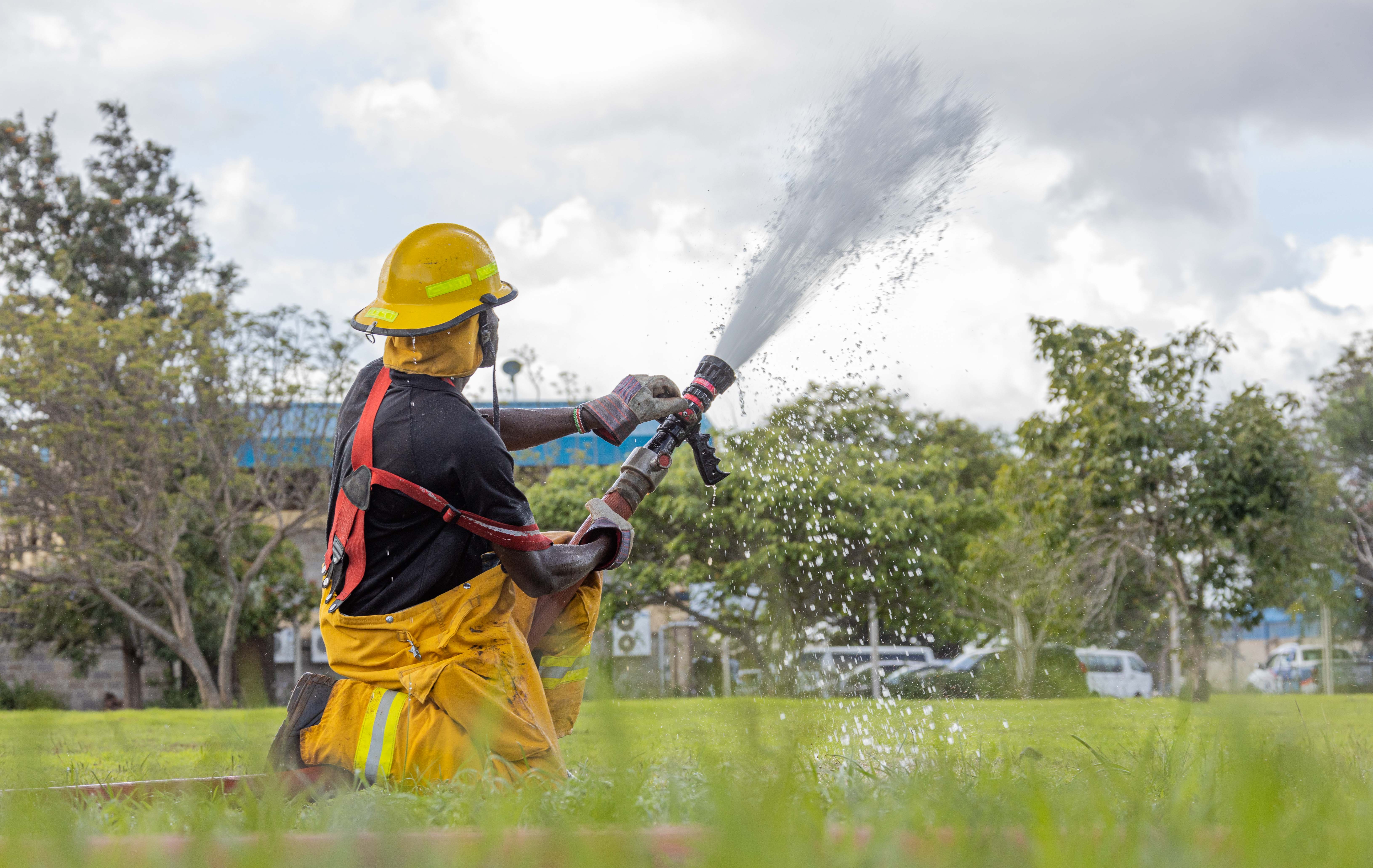 A kneeling firefighter in a yellow helmet sprays a jet of water across a field