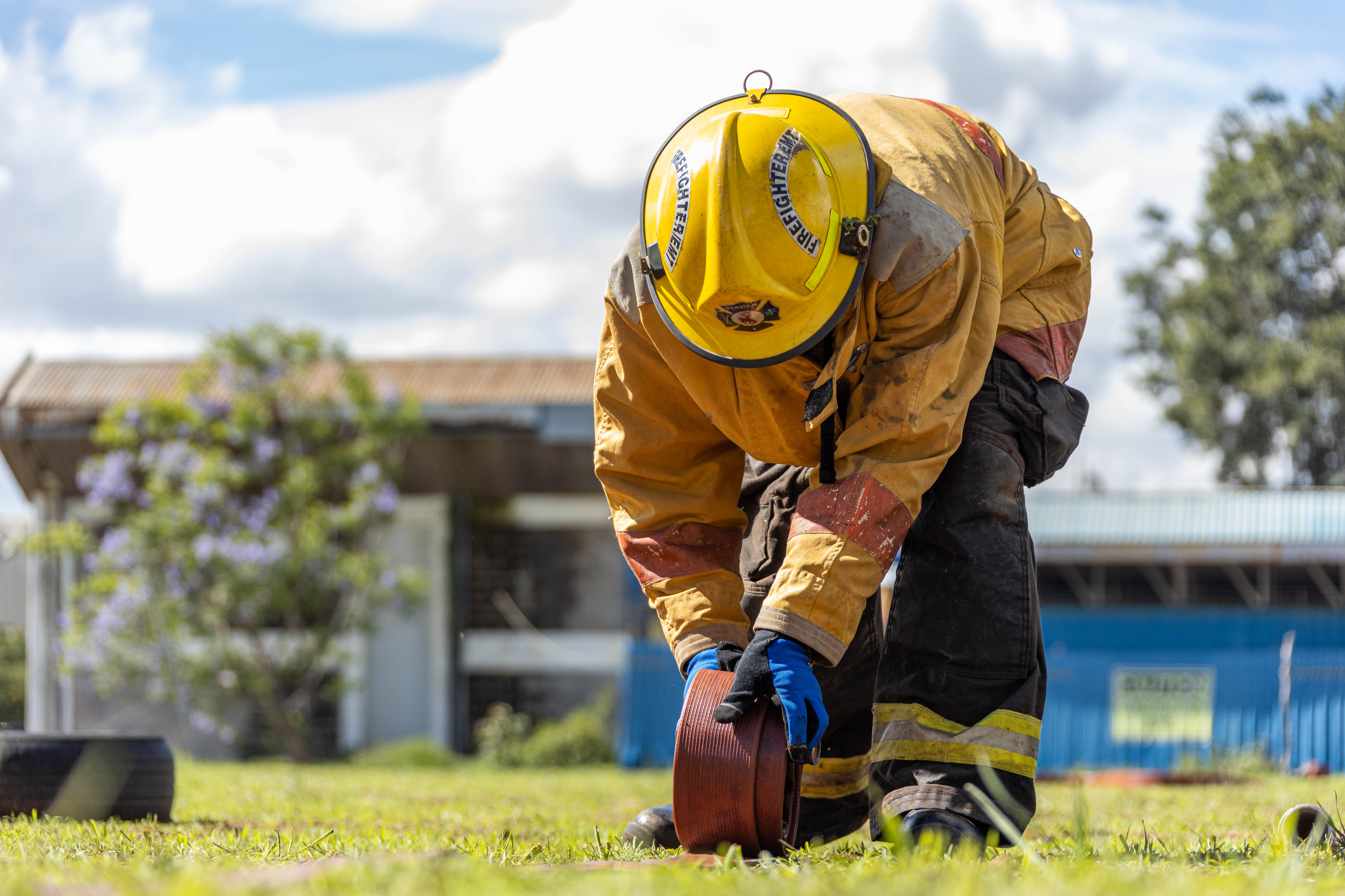 A firefighter in a yellow helmet bends to roll up a red hose on the grass