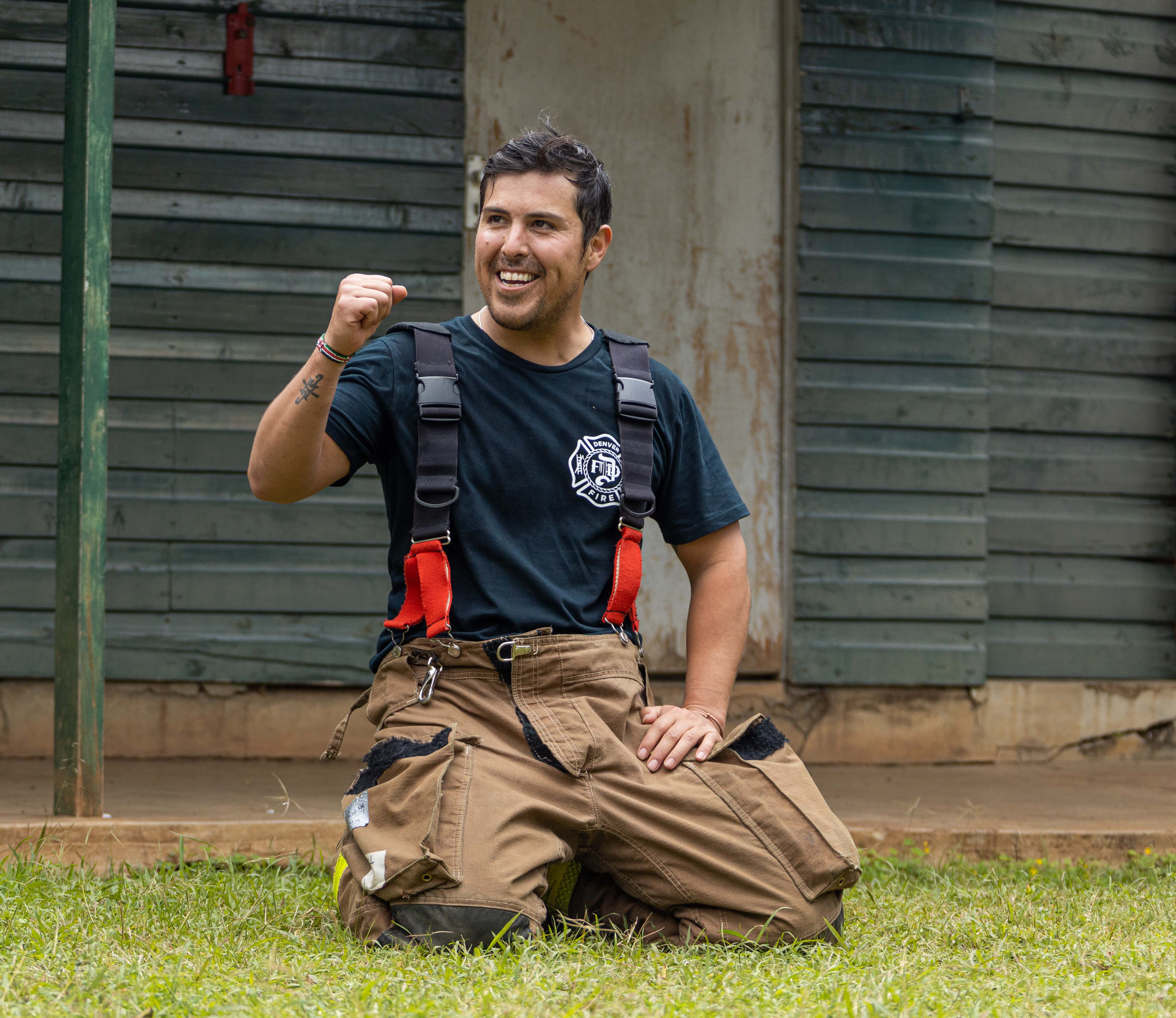 An instructor in fire trousers kneels on the grass, smiling with a raised fist