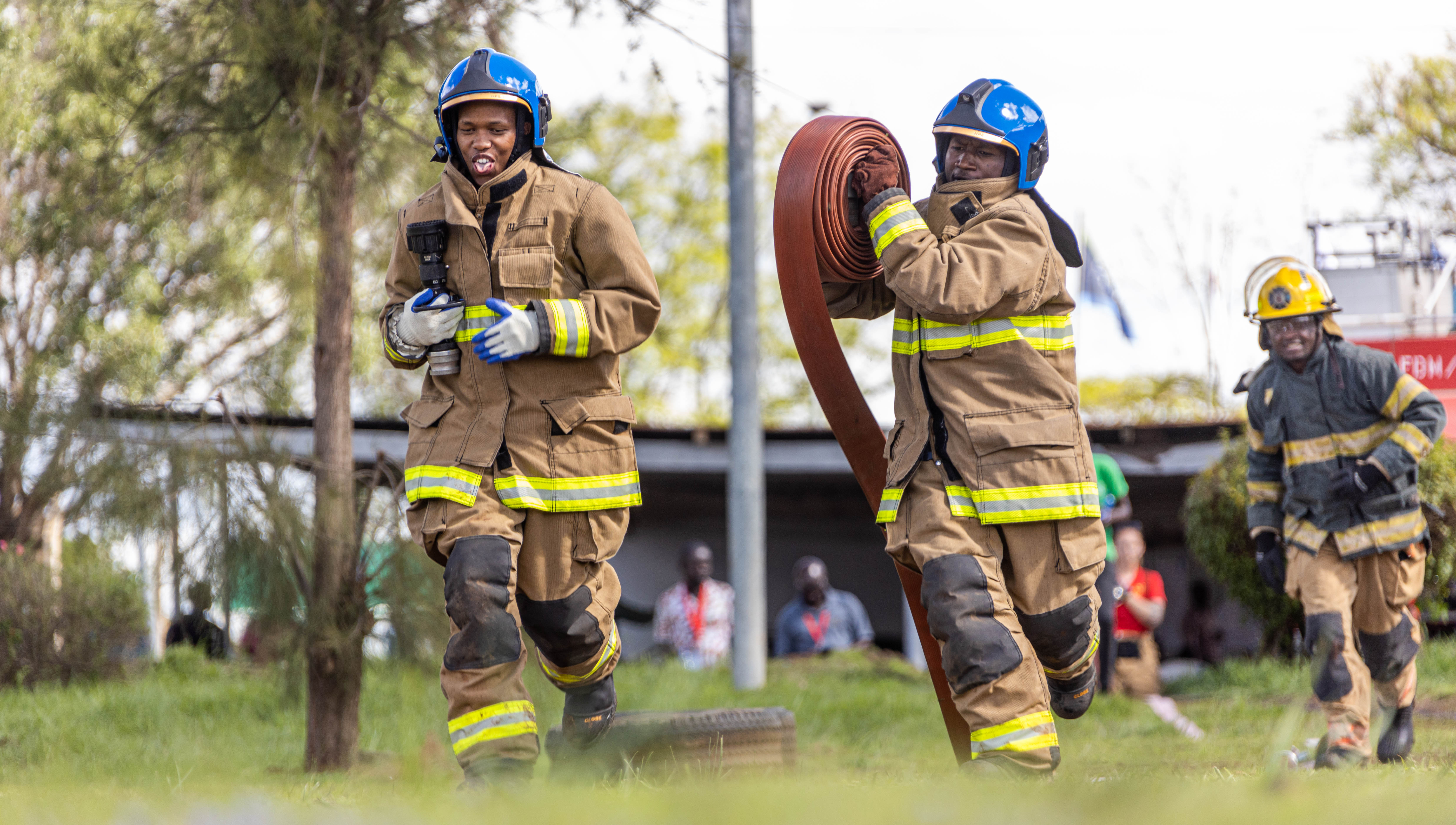 Two firefighters in blue helmets run a timed hose relay while a third follows