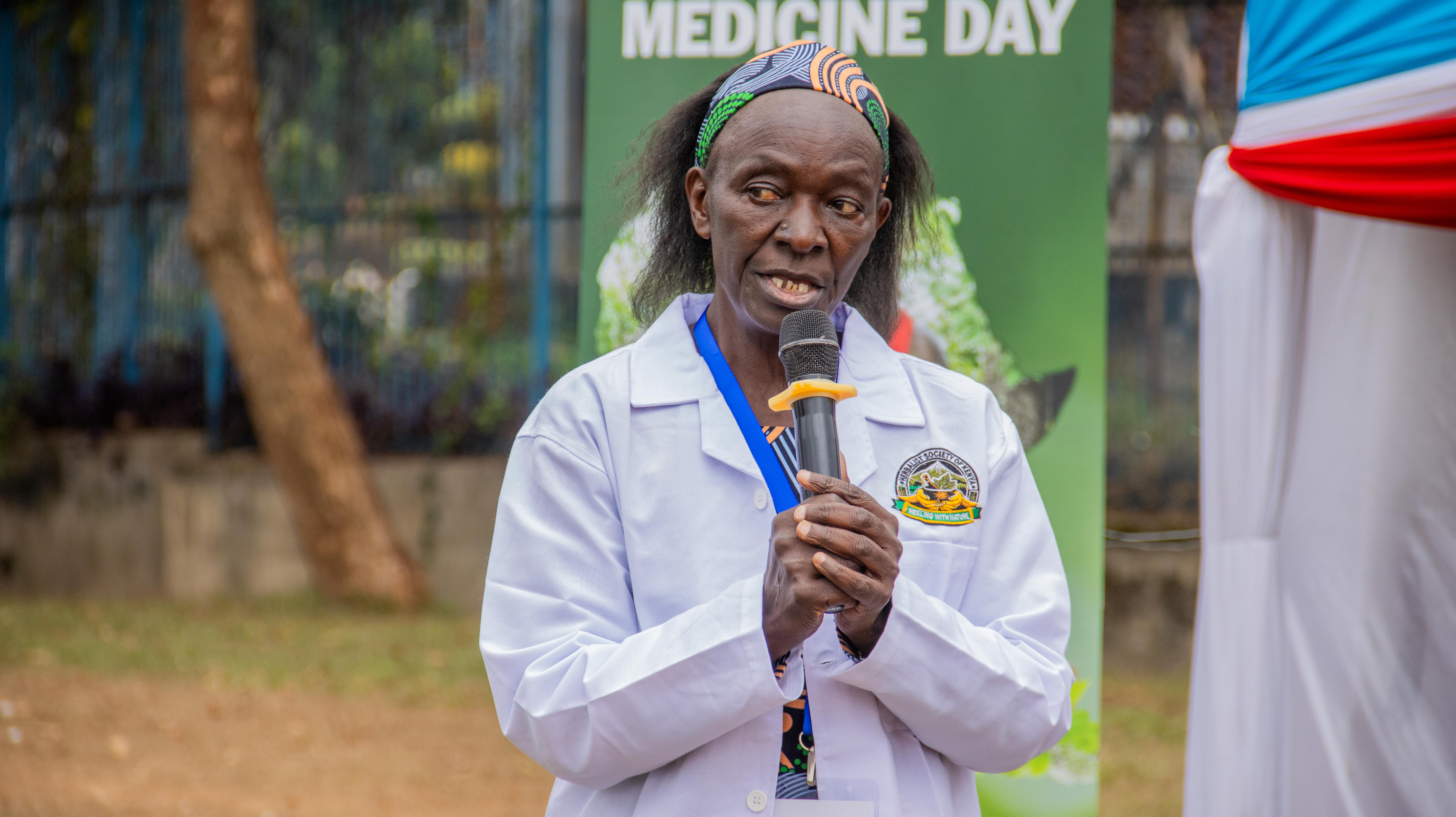 A woman in a white lab coat speaks into a microphone in front of an event banner