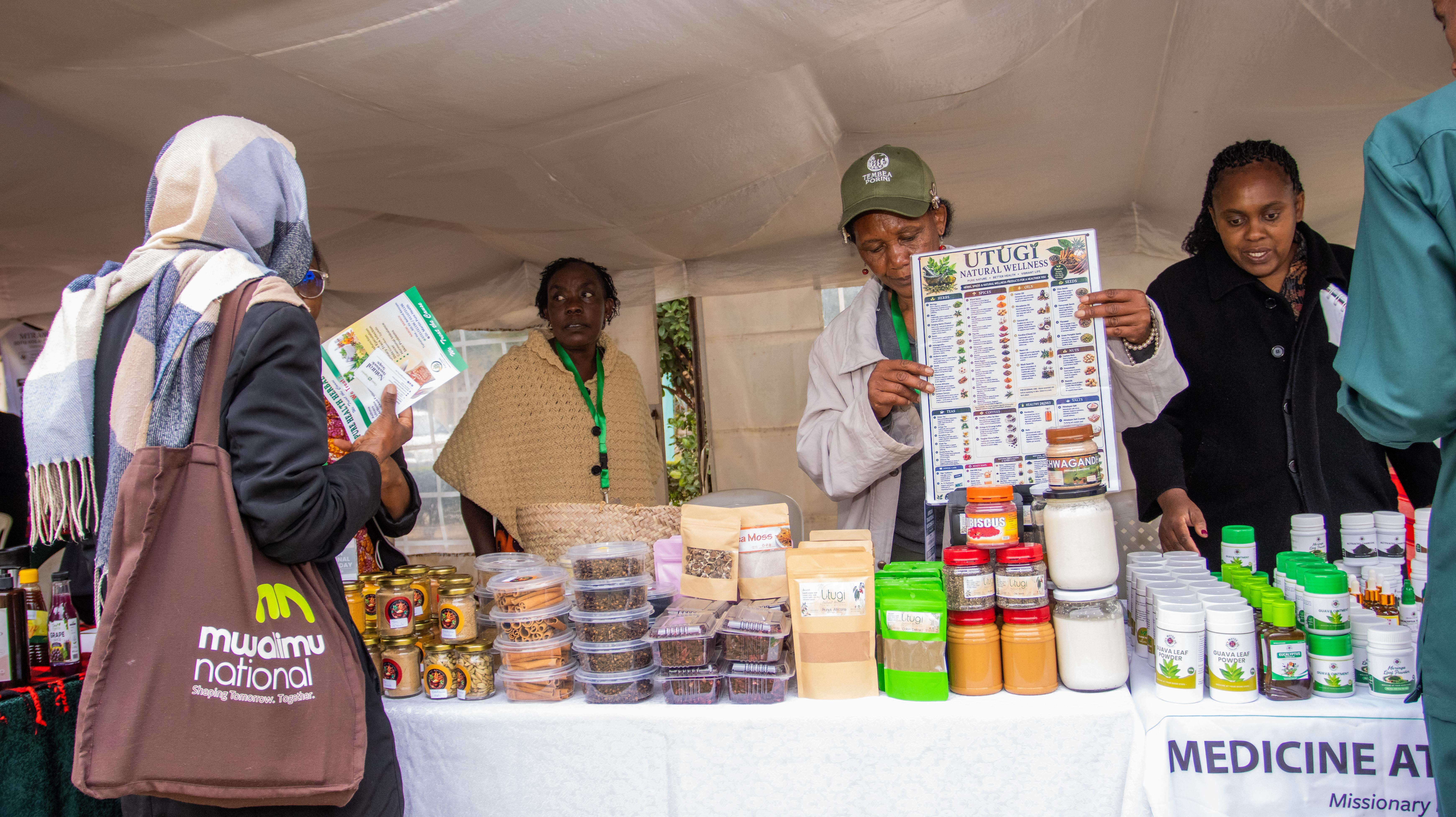 Exhibitors stand behind a table of jars and packets of natural products under a tent