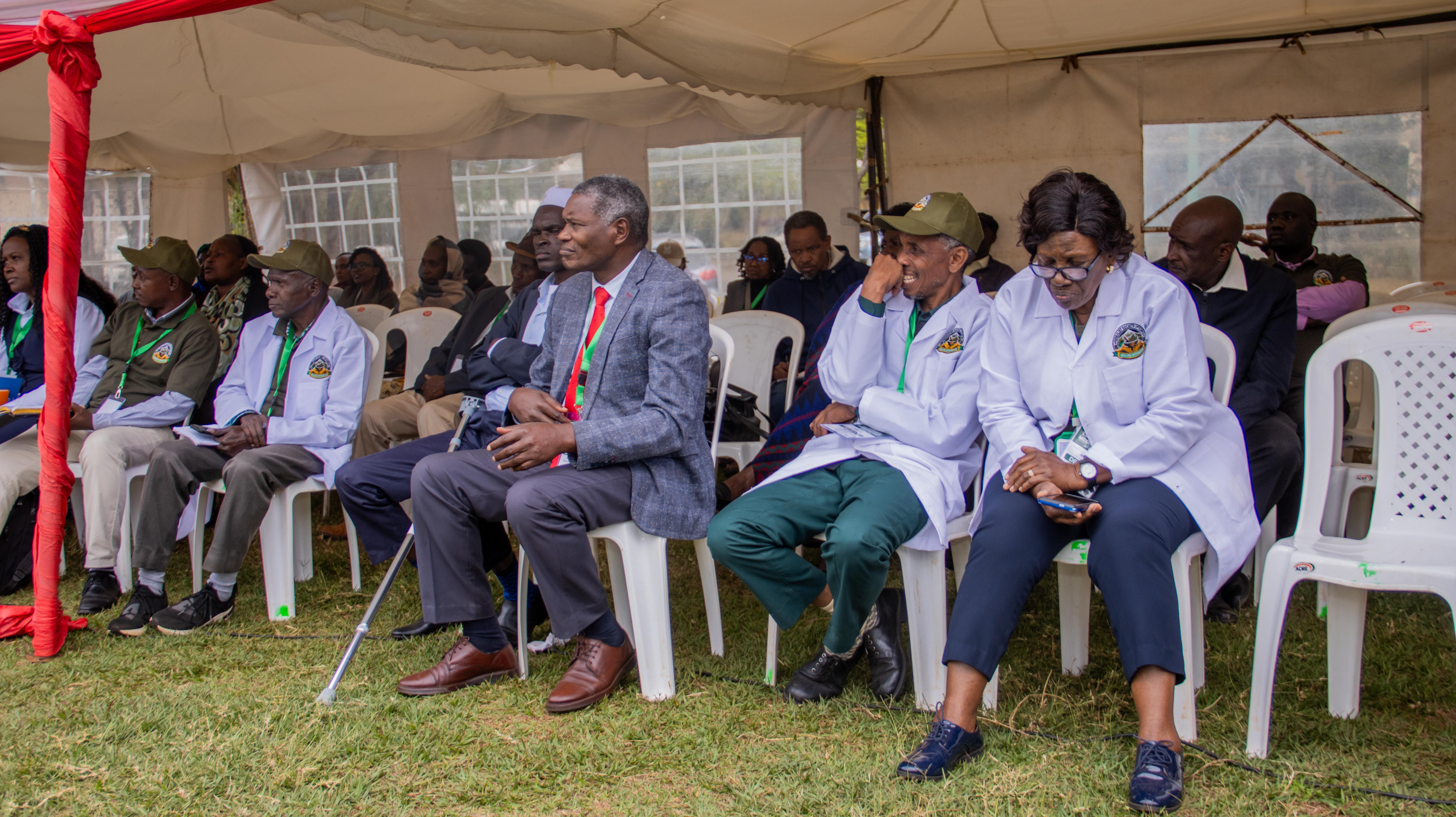 Guests in lab coats and suits sit in rows of chairs under a tent